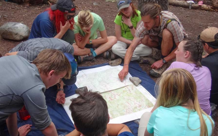 A group of students sit around a map spread out on the ground. One person, who appears to be an instructor, points at the map.